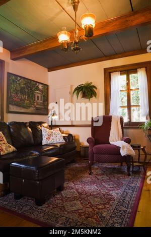 Dark brown leather sofa with footstool and burgundy upholstered armchair in living room inside reconstructed 1840s log home. Stock Photo