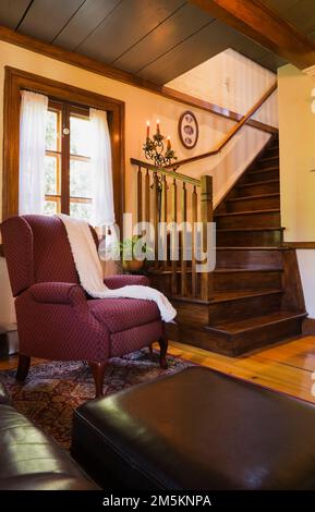 Dark brown leather sofa with footstool and burgundy upholstered armchair next to wooden staircase in living room inside reconstructed 1840s log home. Stock Photo