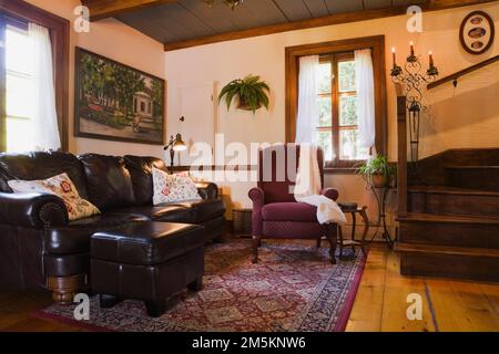 Dark brown leather sofa with footstool and burgundy upholstered armchair next to wooden staircase in living room inside reconstructed 1840s log home. Stock Photo