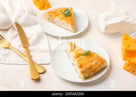 Plates with pieces of delicious cheese pie, parsley leaf, cutlery and ingredients on white table Stock Photo