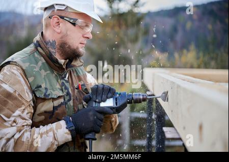Man building wooden frame house. Worker drilling hole by electric drill in timber framing of future house. Builder wearing white helmet and protective goggles. Carpentry concept. Stock Photo