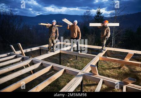 Men workers building wooden frame house on pile foundation. Portrait of three carpenters carrying wooden planks for timber framing. Carpentry concept. Stock Photo