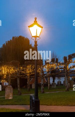 Old-fashioned street lamp lit up at night in Odiham churchyard, Hampshire, England, UK, with Christmas lights on trees Stock Photo