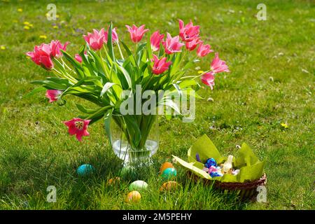 Beautiful pink roses in a vase, colorful Easter eggs, and a basket with chocolates on the grass Stock Photo