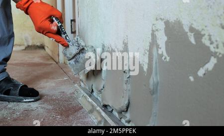 leveling a room wall with a thick layer of plastic putty using a spatula in an orange-gloved hand, applying cement mortar on the surface of the wall Stock Photo