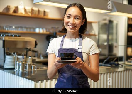 Smiling asian girl barista making coffee, holding cup, wearing cafe uniform apron, standing near counter Stock Photo