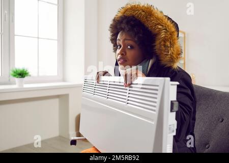 African American woman in winter clothes sitting on couch with electric heater in cold house Stock Photo