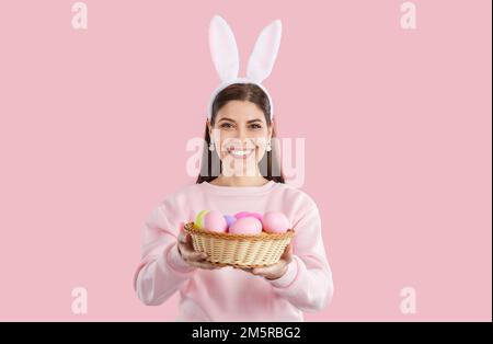 Happy girl in pink sweatshirt with bunny ears giving basket with colorful eggs on pink background. Stock Photo
