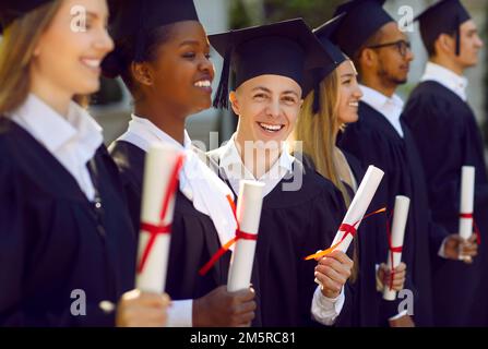 Happy graduate man in graduation gown and cap with diploma posing for photo standing among students. Stock Photo