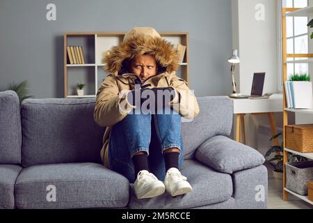 Young frozen indian man sitting in a winter coat on the sofa at home. Heating problems concept. Stock Photo