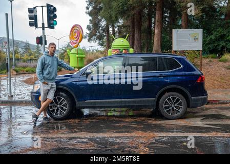 Man leaning on blue car parked on wet street in front of android statues Stock Photo