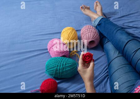 Goiania, Goiás, Brazil – December 30, 2022: A woman sitting on a bed with spools of colored thread around her and a detail of a hand holding a rose. Stock Photo