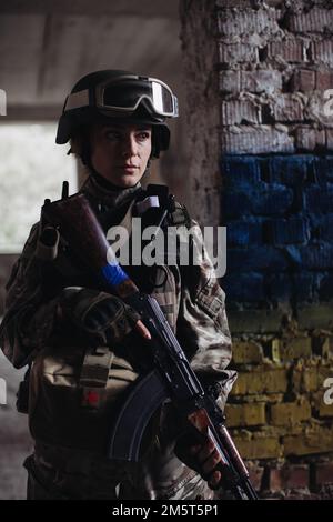 Military female soldier against the background of the flag of Ukraine. The flag of Ukraine is painted on a brick wall, a Ukrainian defender with a wea Stock Photo