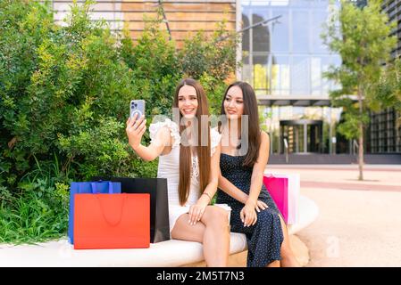 Fashionable girlfriends taking selfie on bench after shopping in boutiques Stock Photo