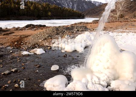 A stream of a small mountain stream flows down onto an ice block of frozen water on the bank of a beautiful river on a winter day. Altai, Siberia, Rus Stock Photo