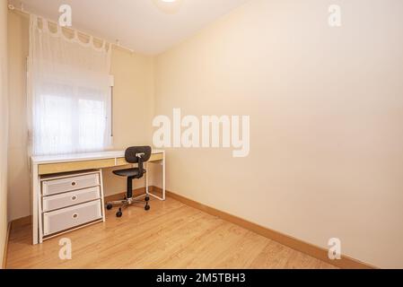 A mostly empty room with a white study table with a chest of drawers and a black study swivel chair with poor back support Stock Photo