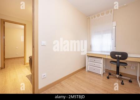 A nearly empty room with a white study table with a chest of drawers and a black swivel study chair with poor back support and access to other rooms w Stock Photo