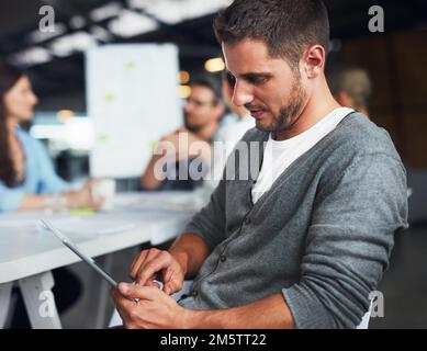 Double checking his digital documents for the meeting. a young man sitting at a table in an office using a digital tablet with colleagues working in Stock Photo