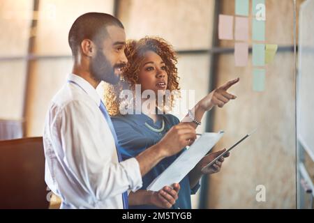 Professionals in action. two young professionals brainstorming with sticky notes on a glass wall in an office. Stock Photo