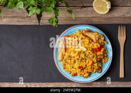 Blue plate with mixed paella, meat and fish, on a gray tablecloth, a wooden fork and half a lemon, on a rustic wooden table with parsley garnish. Stock Photo