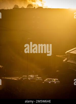 West Yorkshire, UK. 30th Dec, 2022.  Cars drive up a hill near the Shibden valley as the leave Halifax, West Yorkshire, with headlights on and storm clouds and harsh winter sunlight drops below the hill behind silhouetting a farm house. Credit: Windmill Images/Alamy Live News Stock Photo