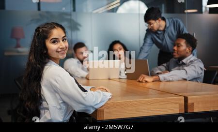 Indian ethnic businesswoman looking at camera posing in office on blurred background of brainstorming diverse office workers multiethnic multiracial Stock Photo