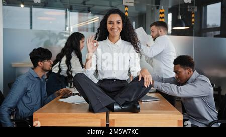 Happy calm smiling businesswoman female leader woman sitting on table in lotus position meditating in office showing ok hand gesture okay sign on Stock Photo