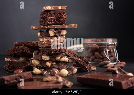 Pieces of broken tasty milk, dark and air chocolate bars with hazelnut  and cocoa beans in glaas jar on black background Stock Photo