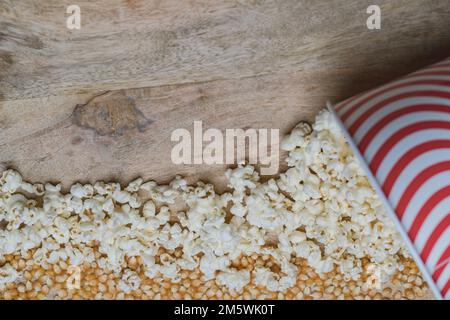 Popcorn (popped) and kernels spilling from a red and white striped container onto wood. Snack food. Stock Photo