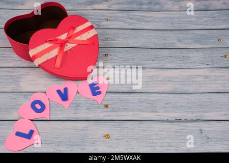Red heart-shaped box on top of a blue wooden table, with some hearts with the word love. Stock Photo