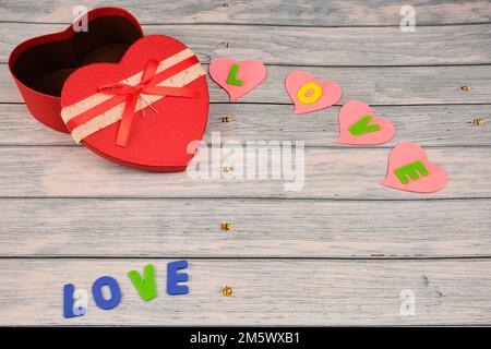 Red heart-shaped box on top of a blue wooden table, with some hearts with the word love. Stock Photo