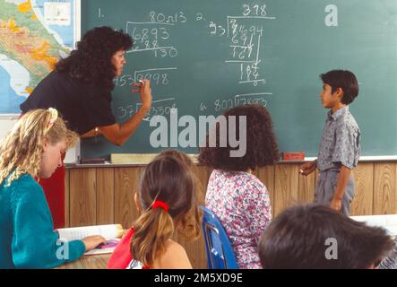 Teacher at the blackboard, writing, math problems. Well, a boy student stands trying to figure out the answers Stock Photo