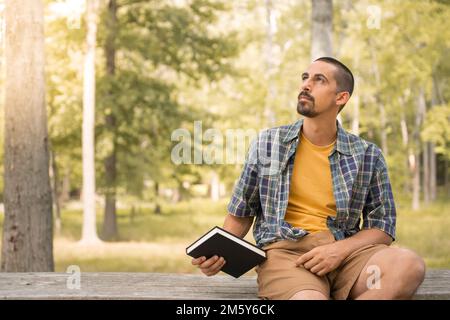 Young man sitting outdoors holding book in a park with trees background - Knowledge faith concept. Stock Photo