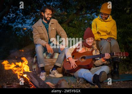 Three happy diverse friends having fun playing music and enjoying bonfire camping in nature at night Stock Photo