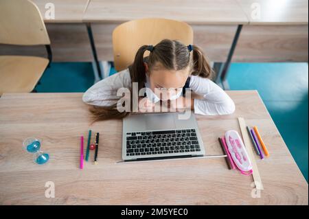 Caucasian girl sits at a desk at school and studies at a laptop. Hourglass on the table. Stock Photo