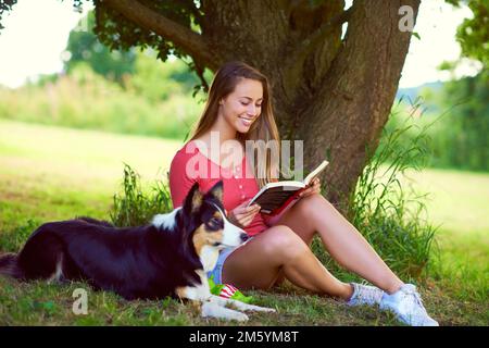 Shes in her happy place. a young woman reading a book while sitting with her dog under a tree. Stock Photo