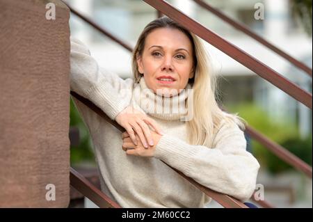 Attractive blond middle-aged woman sitting on a staircase leaning against the railing and looking into the distance Stock Photo