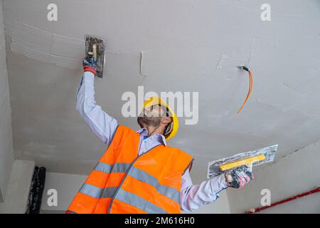 A plasterer in construction wears overalls and a hat. He is plastering a wall with finishing putty using a putty knife. Repair work and construction c Stock Photo