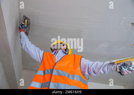 A plasterer in construction wears overalls and a hat. He is plastering a wall with finishing putty using a putty knife. Repair work and construction c Stock Photo