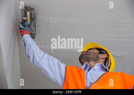 A plasterer in construction wears overalls and a hat. He is plastering a wall with finishing putty using a putty knife. Repair work and construction c Stock Photo