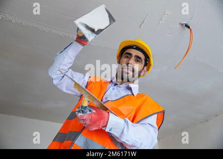 A plasterer in construction wears overalls and a hat. He is plastering a wall with finishing putty using a putty knife. Repair work and construction c Stock Photo