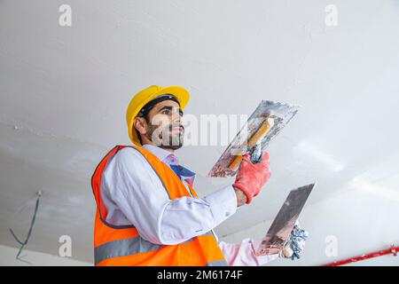 A plasterer in construction wears overalls and a hat. He is plastering a wall with finishing putty using a putty knife. Repair work and construction c Stock Photo