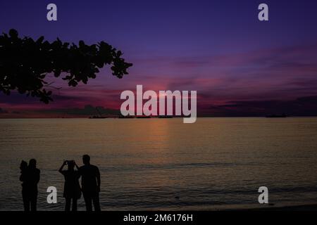 Silhouettes on a beach at sunset in Malaysia, with vibrant purple and pink skies reflecting on calm water. Stock Photo