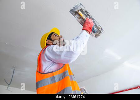 A plasterer in construction wears overalls and a hat. He is plastering a wall with finishing putty using a putty knife. Repair work and construction c Stock Photo