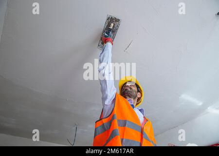 A plasterer in construction wears overalls and a hat. He is plastering a wall with finishing putty using a putty knife. Repair work and construction c Stock Photo