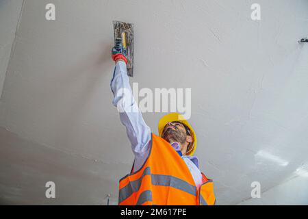A plasterer in construction wears overalls and a hat. He is plastering a wall with finishing putty using a putty knife. Repair work and construction c Stock Photo
