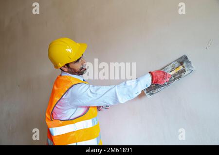 A plasterer in construction wears overalls and a hat. He is plastering a wall with finishing putty using a putty knife. Repair work and construction c Stock Photo