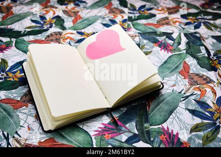 Notebook open to a blank sheet with a pink heart on a sheet, on top of a table with a tropical pattern. Stock Photo