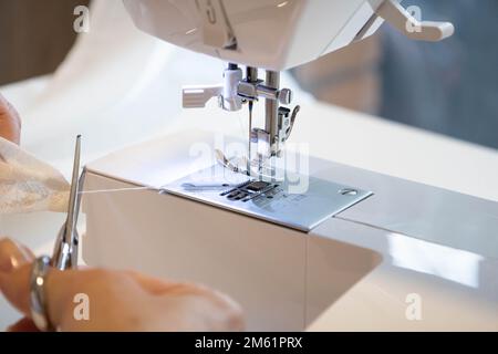 Close up top side view of cropped female hands sewing fabric in manufacturing machine at workplace cutting thread with scissors. Stock Photo