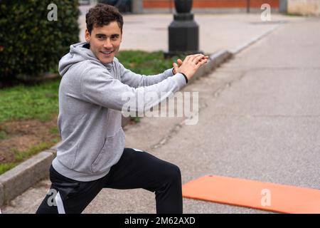Smiling young male athlete in gray hoodie doing lunges while stretching muscles before running in park Stock Photo